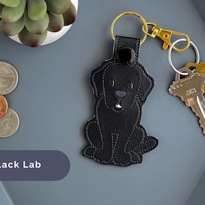 Puede incluir: Llavero de perro Labrador Retriever negro con una correa de cuero negro y un anillo de llave de metal dorado. El llavero est&aacute; sobre una superficie gris con monedas.