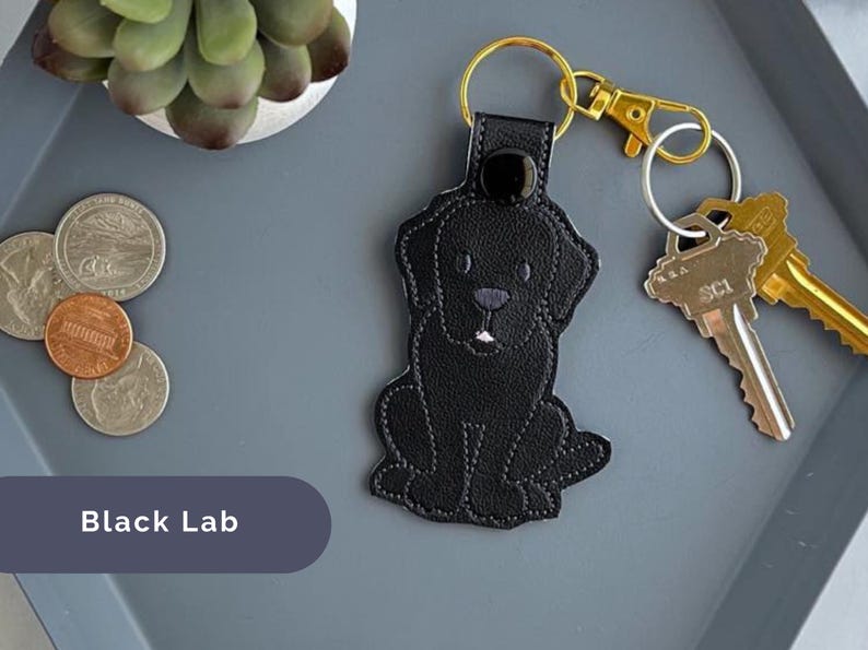 Puede incluir: Llavero de perro Labrador Retriever negro con una correa de cuero negro y un anillo de llave de metal dorado. El llavero est&aacute; sobre una superficie gris con monedas.
