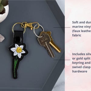 May include: A black leather keychain with an embroidered white daffodil. The keychain includes a silver or gold split keyring and swivel clasp hardware.  The keychain is made of soft and durable marine vinyl (faux leather) fabric.