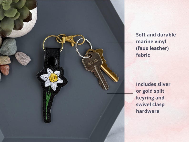 May include: A black leather keychain with an embroidered white daffodil. The keychain includes a silver or gold split keyring and swivel clasp hardware.  The keychain is made of soft and durable marine vinyl (faux leather) fabric.