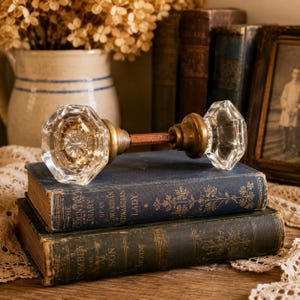 May include: A vintage door knob set featuring two clear, multi-faceted glass knobs connected by a central metal spindle. The glass knobs have a geometric, octagonal shape with a starburst pattern at the center, attached to aged brass-toned metal bases.