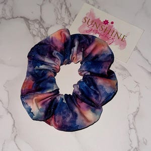 May include: A scrunchie with a galaxy print in shades of blue, purple, pink, and orange. The fabric is gathered and elasticized. A small card with the text "Sunshine Scrunchies" is visible.