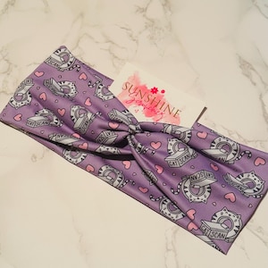 Puede incluir: Una diadema lavanda con un patrón repetido de gatos de dibujos animados dentro de máquinas CAT SCAN, con corazones rosas. La diadema tiene un diseño de nudo retorcido. Una pequeña tarjeta con el texto "SUNSHINE" está en la diadema.