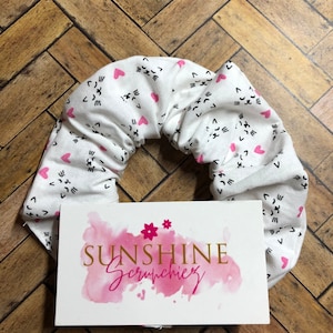 May include: White fabric scrunchie with a pink heart and black cat face pattern. The scrunchie is on a wooden surface with a pink and gold business card that says "Sunshine Scrunchies".