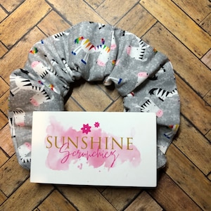 May include: Grey scrunchie with a zebra and rainbow zebra print. The scrunchie is on a wooden background. The text "Sunshine Scrunchies" is on a white card with a pink watercolour background.