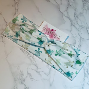 Op de afbeelding: Een wit hoofdband met een groene en blauwe waterverf schildpadprint. Het hoofdband is gedraaid en heeft een klein visitekaartje met de tekst "Sunshine Jewelry".