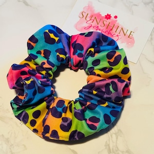 May include: A colorful scrunchie with a rainbow leopard print pattern. The scrunchie features a vibrant mix of colors, including blue, pink, yellow, and green, with dark purple spots. The scrunchie is next to a card that says "Sunshine Scrunchies".