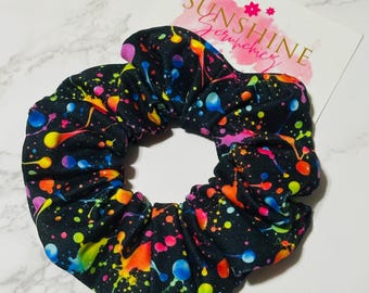 Könnte beinhalten: Ein schwarzes Stoff-Haargummi mit einem lebendigen, mehrfarbigen Farbspritzer-Muster in Neon-Pink, Gelb, Orange, Blau und Grün. Dahinter befindet sich eine weiße Präsentationskarte mit dem Text SUNSHINE Scrunchies.