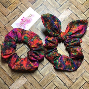 Puede incluir: Dos scrunchies de pelo coloridos con un diseño de pintura salpicada. Un scrunchie está atado en un lazo.