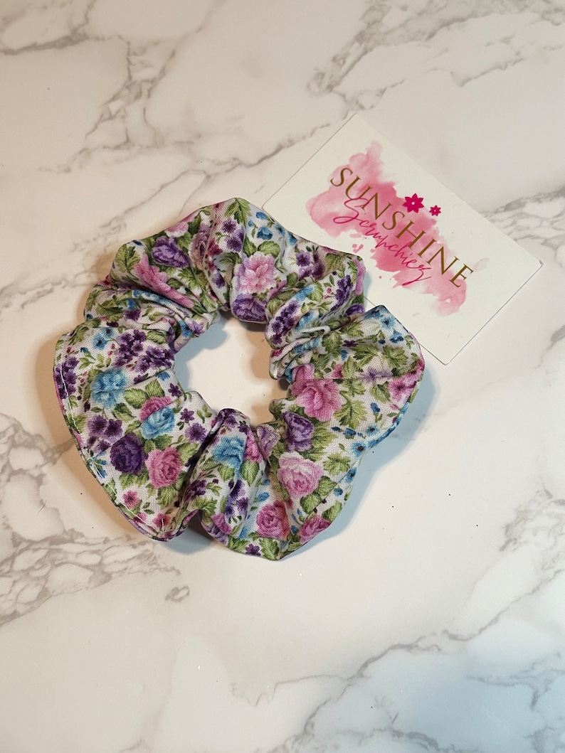 K&ouml;nnte beinhalten: Ein wei&szlig;er Haargummi mit einem Blumenmuster mit rosa, lila und blauen Blumen. Der Haargummi ist aus Stoff und ist zu einem Knoten gebunden.