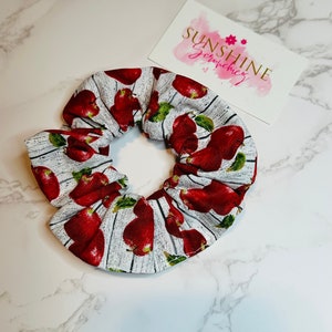 May include: A white scrunchie with a pattern of red pears and green leaves. The scrunchie is on a white and gray surface. A card with the text "SUNSHINE Scrunchiez" is also visible.