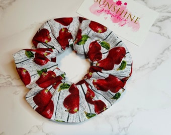 May include: A white scrunchie with a pattern of red pears and green leaves. The scrunchie is on a white and gray surface. A card with the text "SUNSHINE Scrunchiez" is also visible.