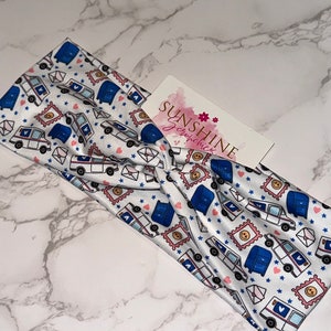 May include: A white headband with a blue and white mail truck pattern. The headband is tied in a knot and has a white background with pink hearts, blue stars, and white envelopes.