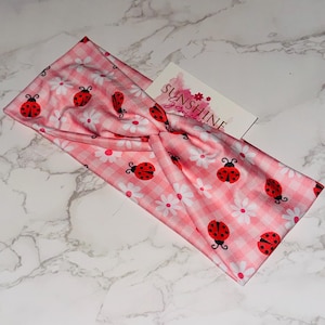 May include: Pink and white gingham headband with a pattern of red ladybugs and white daisies. The headband has a twisted knot design. A small white card with the text "Sunshine Jewels" is visible.