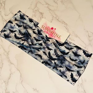 Op de afbeelding: Een brede stoffen hoofdband met een patroon van zwarte vleermuizen op een bewolkte blauwe en grijze achtergrond. De hoofdband is gevouwen en gedraaid. Een kleine witte kaart met de tekst "SUNSHINE Scrunchies" is zichtbaar.
