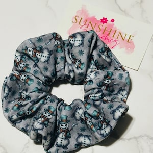 May include: A gray fabric hair scrunchie featuring a repeating pattern of snowmen wearing teal top hats and orange scarves, interspersed with small snowflakes. A white rectangular card with the text SUNSHINE scrunchiez is visible behind it.