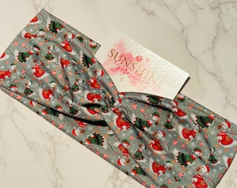 May include: A gray fabric headband with a repeating pattern of snowmen wearing red and green outfits. The headband has a twisted knot design. A small card with the text "SUNSHINE" is visible.
