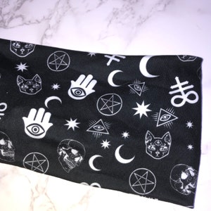 Op de afbeelding: Zwarte stof met een wit, herhalend patroon van verschillende occulte symbolen, waaronder een pentagram, een oog, een halve maan, een schedel en een kat met drie ogen.