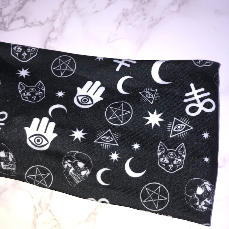 Op de afbeelding: Zwarte stof met een wit, herhalend patroon van verschillende occulte symbolen, waaronder een pentagram, een oog, een halve maan, een schedel en een kat met drie ogen.