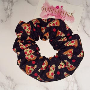 May include: A black fabric hair scrunchie featuring a repeating pattern of pizza slices with red pepperoni and green peppers. A white rectangular card with the text Sunshine Scrunchies in pink and gold lettering is positioned behind the scrunchie.