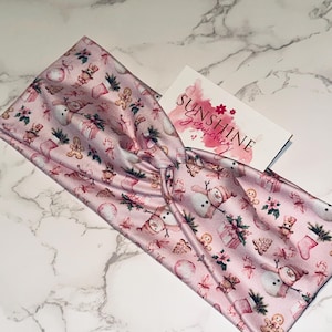 May include: A pink headband with a Christmas-themed pattern featuring snowmen, gingerbread men, and gift boxes. The headband is twisted at the top and rests on a marble surface. A business card with the text "Sunshine Scrunchies" is also present.