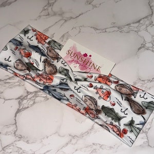 May include: A white headband with a pattern of red and orange fish, blue waves, and fishing hooks. The headband is made of a soft, stretchy fabric and is perfect for adding a touch of whimsy to any outfit.