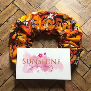May include: A Halloween-themed scrunchie featuring a repeating pattern of orange jack-o'-lanterns and black cats on a black background. The scrunchie is positioned over a white card with the text "SUNSHINE Scrunchiez" in gold lettering.