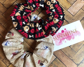 May include: Two fabric scrunchies with dog-themed patterns. One scrunchie is black with red and white accents and features the words "Woof" and "Ruff". The other scrunchie is beige with a pattern of dogs wearing sailor hats. The scrunchies are on a wooden surface.