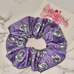 May include: A purple fabric hair scrunchie featuring a repeating pattern of cartoon cats and small pink hearts. The cats are depicted with the text CAT SCAN. A white rectangular card with the text SUNSHINE Scrunchies is positioned nearby.