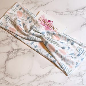 Puede incluir: Una diadema blanca con un patrón azul claro y rosa que presenta la palabra "MOTIVATE" e imágenes de pesas, una escalera y una nube. La diadema está retorcida y tiene una etiqueta blanca con el texto "SUNSHINE Scrunchies" en rosa.