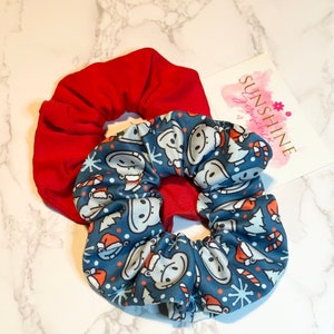 May include: Two scrunchies. One is solid red, the other is patterned with a blue background and a repeating design of cartoon hippos wearing Santa hats, Christmas trees, and candy canes. A card with the text "Sunshine Scrunchies" is in the background.