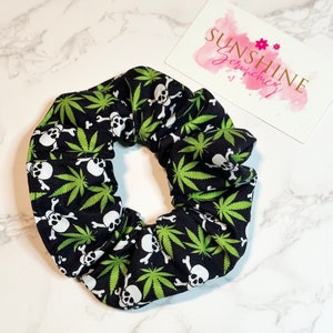May include: Black scrunchie with a repeating pattern of white skulls, crossbones, and green cannabis leaves. The scrunchie is on a white marble surface. A card with the text "SUNSHINE SCRUNCHIES" is in the background.