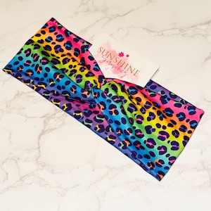 Può includere: Una fascia colorata con stampa leopardata arcobaleno. La fascia presenta un design a nodo attorcigliato e un'etichetta con la scritta "Sunshine Jewelry". I colori vivaci includono rosa, giallo, blu e viola.