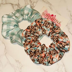 May include: Two scrunchies. One is light green and white gingham. The other is patterned with sloths and flowers on a teal background. A card with the word "SUNSHINE" is in the background.