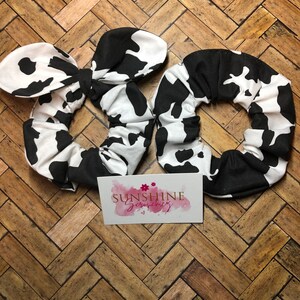 May include: Two black and white cow print scrunchies. One scrunchie is tied in a bow. The scrunchies are on a wooden surface. The text "Sunshine Scrunchies" is on a card in the background.