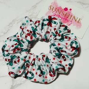 May include: A white fabric hair scrunchie featuring a repeating pattern of red and teal candy canes, red bows, and small red and teal stars. A white rectangular card with the text SUNSHINE Scrunchies in gold and pink script is visible.