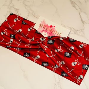 May include: A red fabric headband with a pirate-themed pattern. The design includes white skulls, crossed swords, and pirate flags. The headband has a twisted knot design and a small card with the text "SUNSHINE scrunchies".