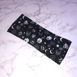 Op de afbeelding: Zwart hoofdband met een wit patroon van verschillende occulte symbolen, waaronder pentagrammen, manen, ogen, schedels en katten.