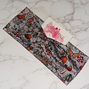 Peut inclure: Bandeau gris avec un motif fantaisiste de champignons, papillons, coccinelles et fleurs. Le bandeau a un design torsadé et présente le texte "SUNSHINE Scrunchies" sur une petite étiquette.