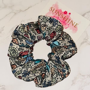 Puede incluir: Una goma para el pelo con un estampado de criaturas marinas de colores sobre un fondo gris claro. La goma para el pelo muestra estrellas de mar, conchas y corales. Junto a ella hay una tarjeta con el texto "SUNSHINE Scrunchies" en rosa.