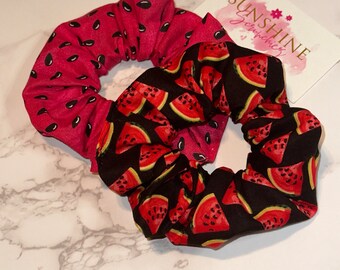 May include: Two fabric scrunchies, one with a watermelon print and one with a pink background and black watermelon seeds. The scrunchies are on a white marble surface. The text "SUNSHINE Scrunchies" is on a white tag.