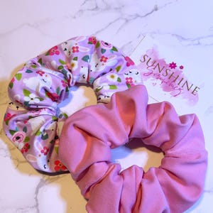 May include: Two hair scrunchies, one with a pink floral pattern featuring bunnies and the other a solid pink color. The scrunchies are on a white marble surface with a business card that reads "Sunshine Jewelry".