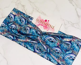 Puede incluir: Una diadema azul con un estampado de delfines. La tela presenta un diseño repetido de delfines sobre un fondo similar al mar con tonos azules, morados y blancos. También se ve una pequeña tarjeta con el texto "Sunshine Scrunchies".