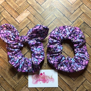 May include: Two purple and pink scrunchies with a mushroom print. The scrunchies are on a wooden surface. The text "Sunshine Scrunchies" is on a white card.