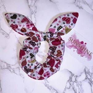 Può includere: Un elastico per capelli bianco con un motivo a cuori rosa, rosso, nero e oro. L'elastico è legato a forma di fiocco. Il testo "Sunshine Jewelry" è visibile sullo sfondo.