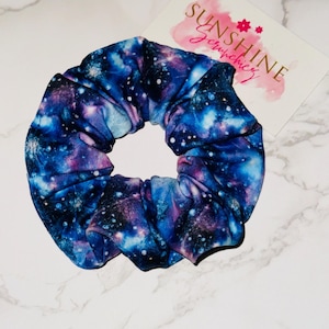 May include: A fabric scrunchie with a galaxy print in shades of blue, purple, and white. The scrunchie is circular and gathered, with a soft, flowing appearance. A card with the text "SUNSHINE Scrunchies" is in the background.