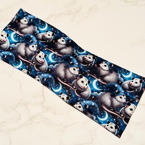 May include: A dark blue headband featuring a repeating pattern of opossums wearing wizard hats, set against a starry night sky with crescent moons. The opossums are gray and white.
