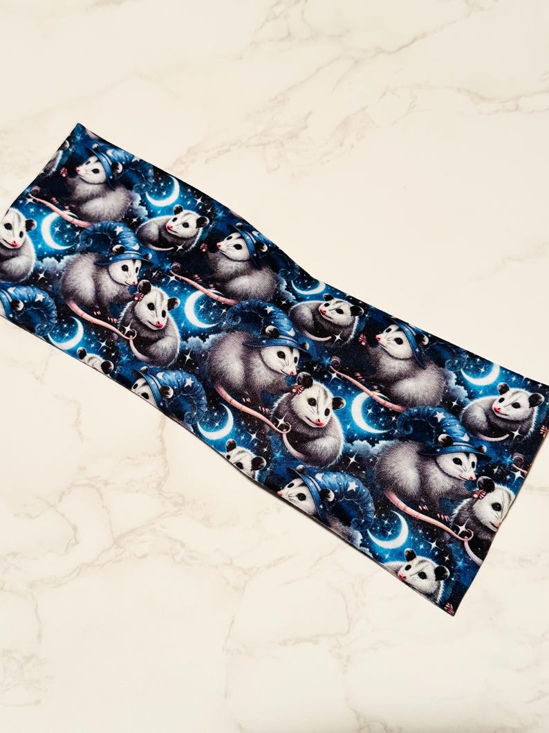 May include: A dark blue headband featuring a repeating pattern of opossums wearing wizard hats, set against a starry night sky with crescent moons. The opossums are gray and white.