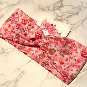 May include: A pink headband with a repeating pattern of desserts, including jars of jam, cupcakes, and macarons. The headband has a twisted knot design. A white card with the text "SUNSHINE scrunchies" is also visible.