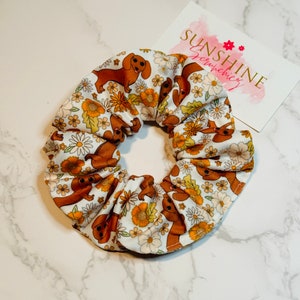 May include: A white scrunchie with a pattern of brown dachshunds and yellow flowers. The scrunchie is made of fabric and has a gathered look. The scrunchie is on a white marble surface. The text "Sunshine Scrunchies" is on a pink and white card behind the scrunchie.